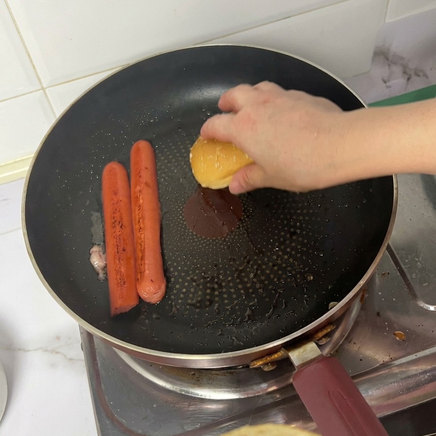 A person placing the top of a sesame seed burger bun face down in a pan next to two browning hot dog sausages.