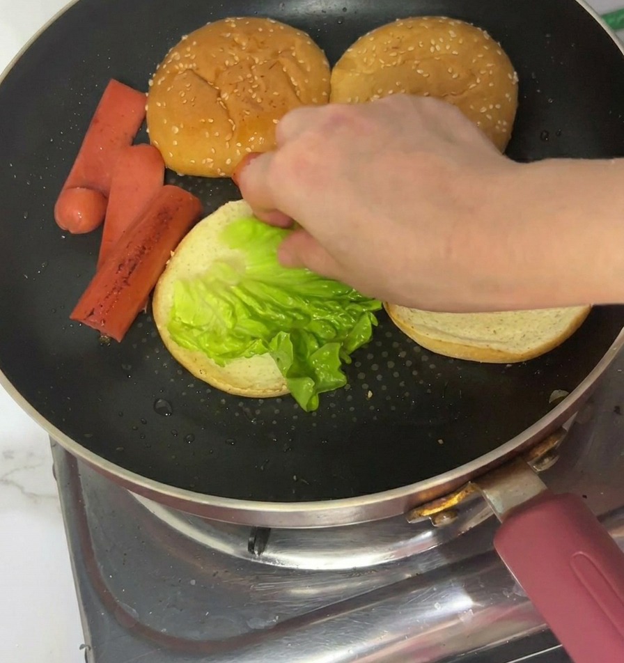 A hand placing a bright green lettuce leaf onto the toasted side of a burger bun sitting in a pan.