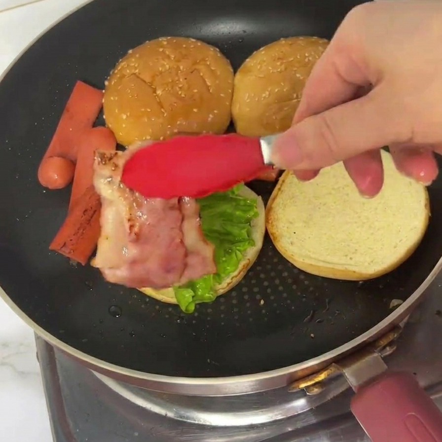 Red tongs placing a strip of cooked, wavy bacon onto a burger bun already topped with lettuce.