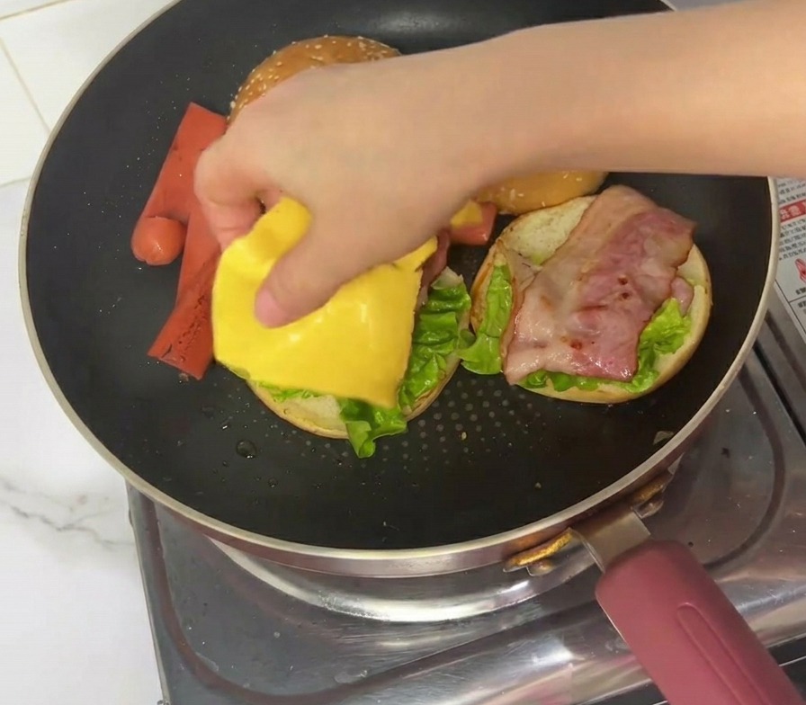 A hand placing a square of yellow processed cheese onto a burger assembly in a frying pan.