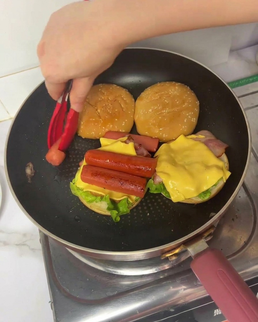 A hand using red tongs to place a folded piece of cooked egg onto a burger layered with lettuce, bacon, cheese, and two hot dog sausages in a frying pan.