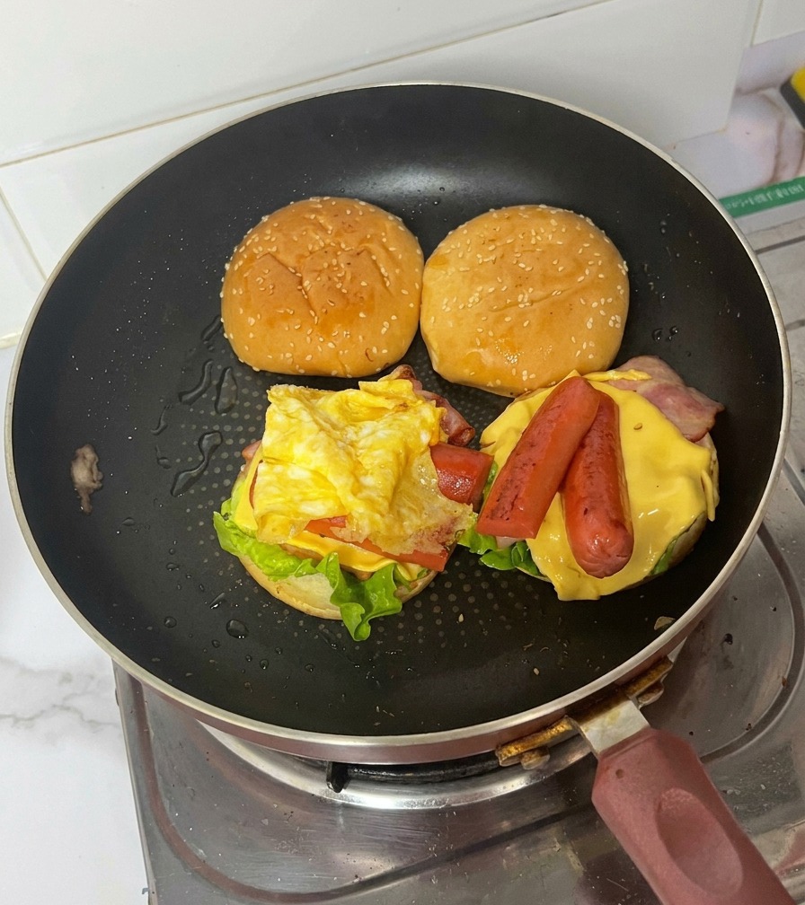 A burger assembly in a frying pan showing a folded fried egg placed over grilled sausages, cheese, bacon, and lettuce.