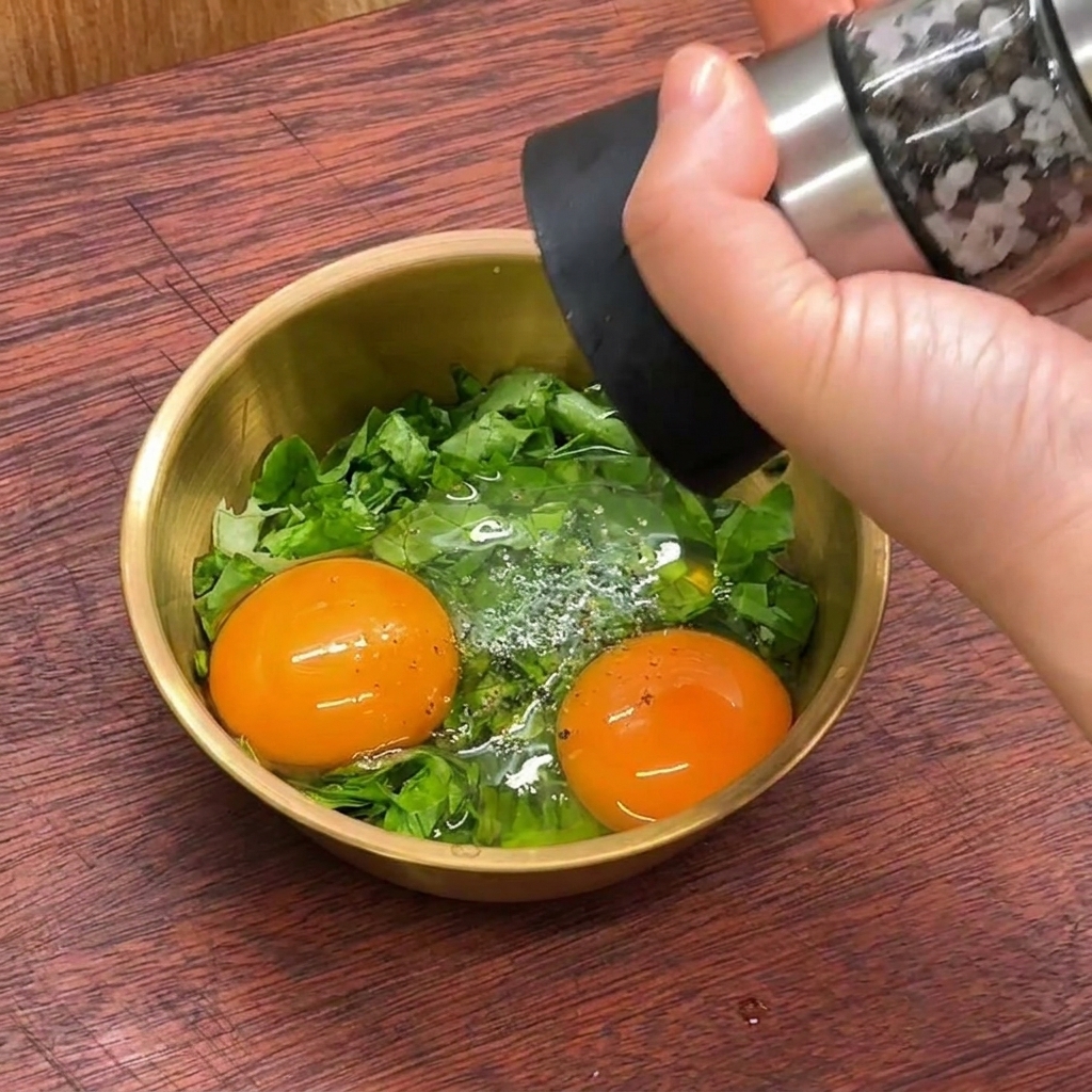 A hand using a pepper mill to season two raw egg yolks and shredded lettuce in a small golden bowl.