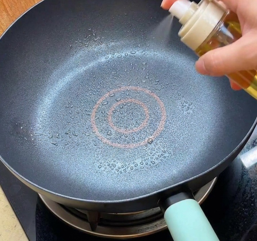 A person using a white spray bottle to mist oil into a clean black non-stick frying pan.