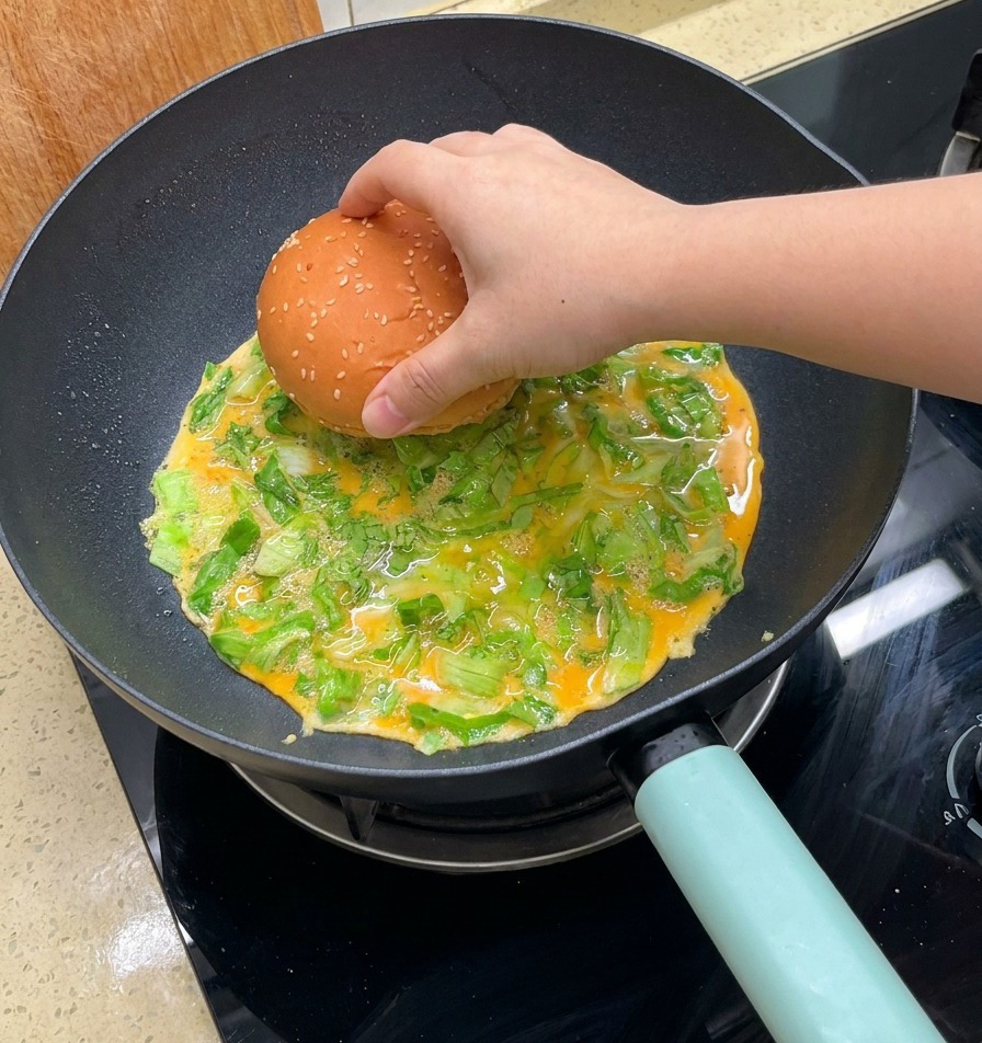 A hand placing a sesame-topped burger bun onto a cooking omelet with shredded lettuce in a pan.