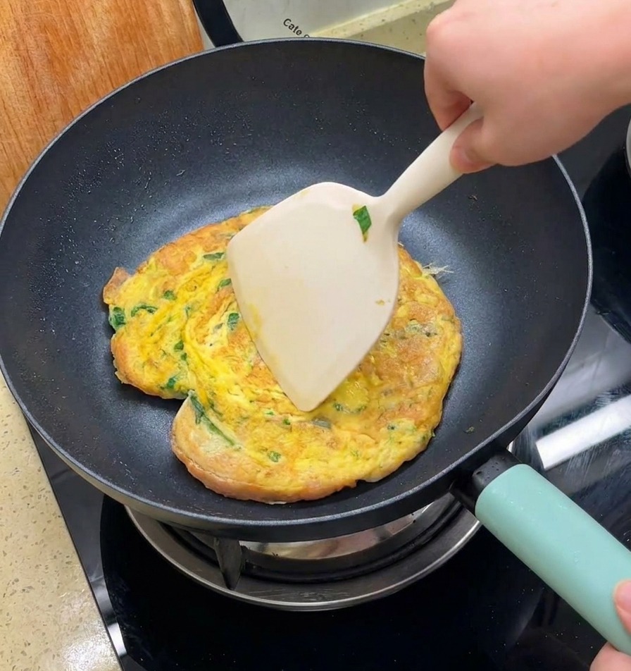 A hand using a large spatula to flip a golden brown egg omelet containing shredded greens and burger buns in a black non stick pan.