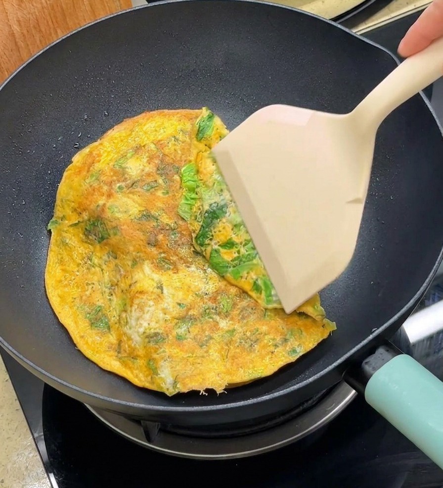 A spatula folding the edges of a round cooked egg sheet over two burger buns in a non-stick pan.