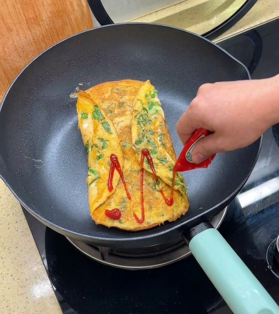 Red ketchup being squeezed from a small packet onto a golden-brown folded egg patty in a frying pan.
