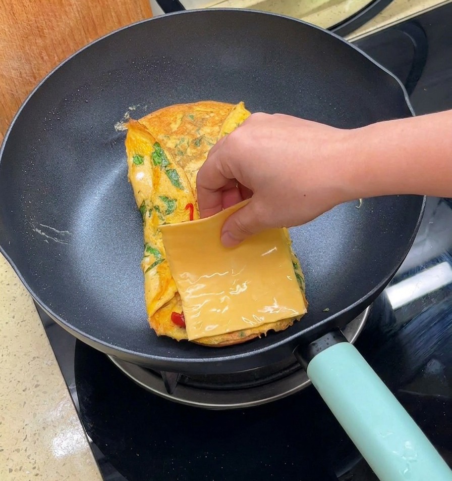 A hand placing a square slice of yellow cheese onto a folded egg sheet in a pan.