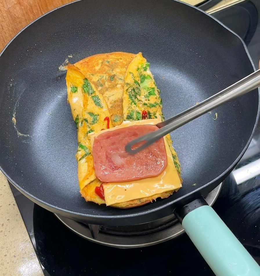 Metal tongs placing a slice of cooked bacon onto cheese on top of a folded omelet in a pan.