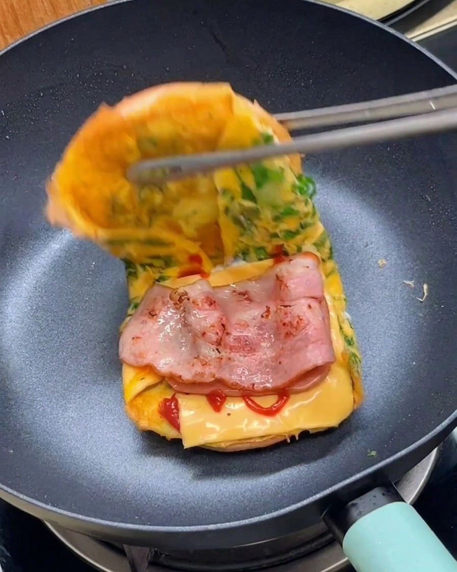 Tongs folding over the edge of a lettuce-filled egg omelet to complete a breakfast burger sandwich in a pan.
