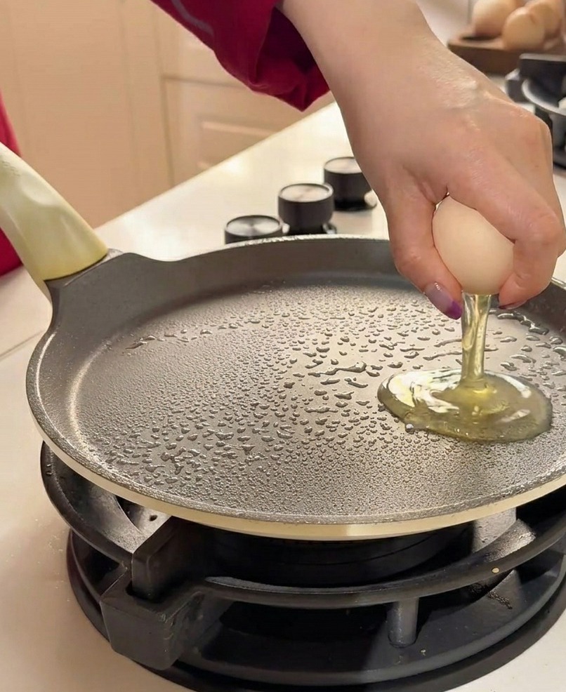 Two raw eggs being cracked onto a greased flat frying pan by hand.