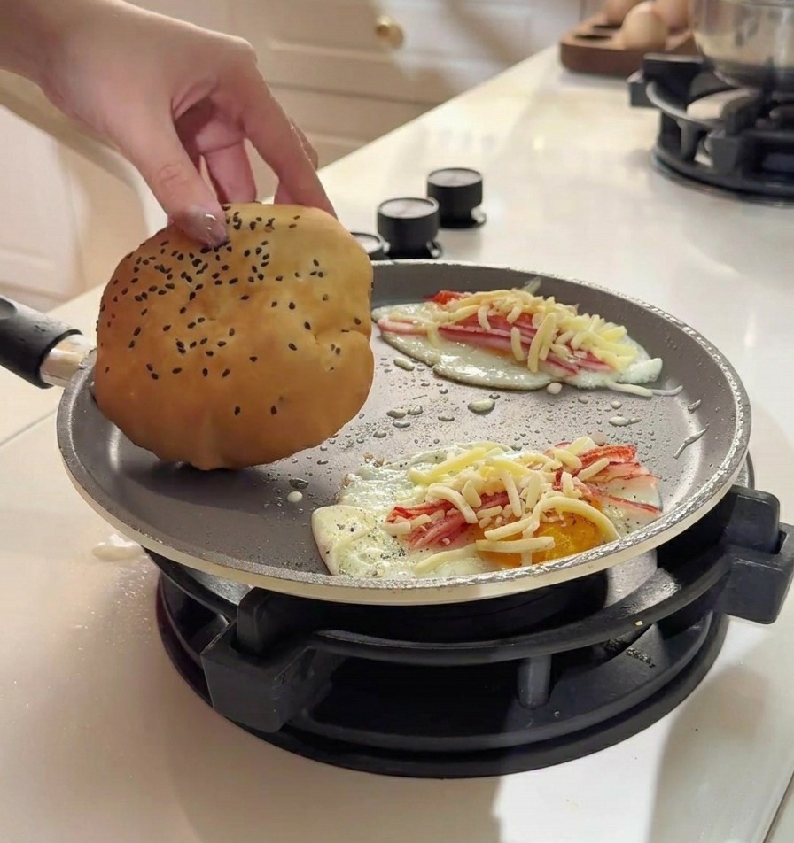 A hand placing a soft, golden-brown sesame bun onto a fried egg topped with crab sticks and melted cheese in a pan.