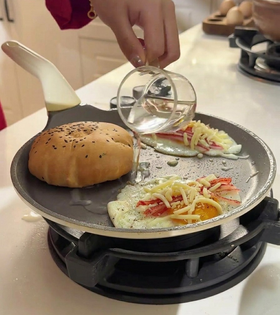 Pouring water from a glass cup into a pan with a burger bun and eggs to create steam.
