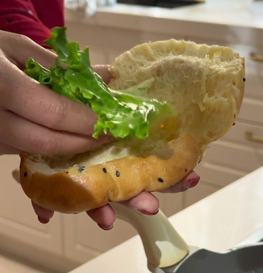 A hand placing a vibrant green leaf of curly lettuce into a soft, sliced butter bun speckled with black sesame seeds.
