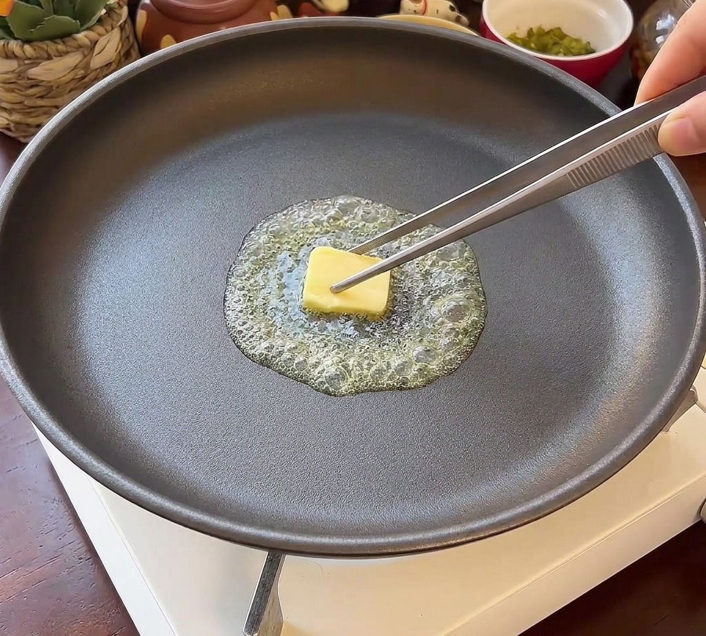 Two round burger bun halves placed face down in a dark frying pan with shimmering melted butter.