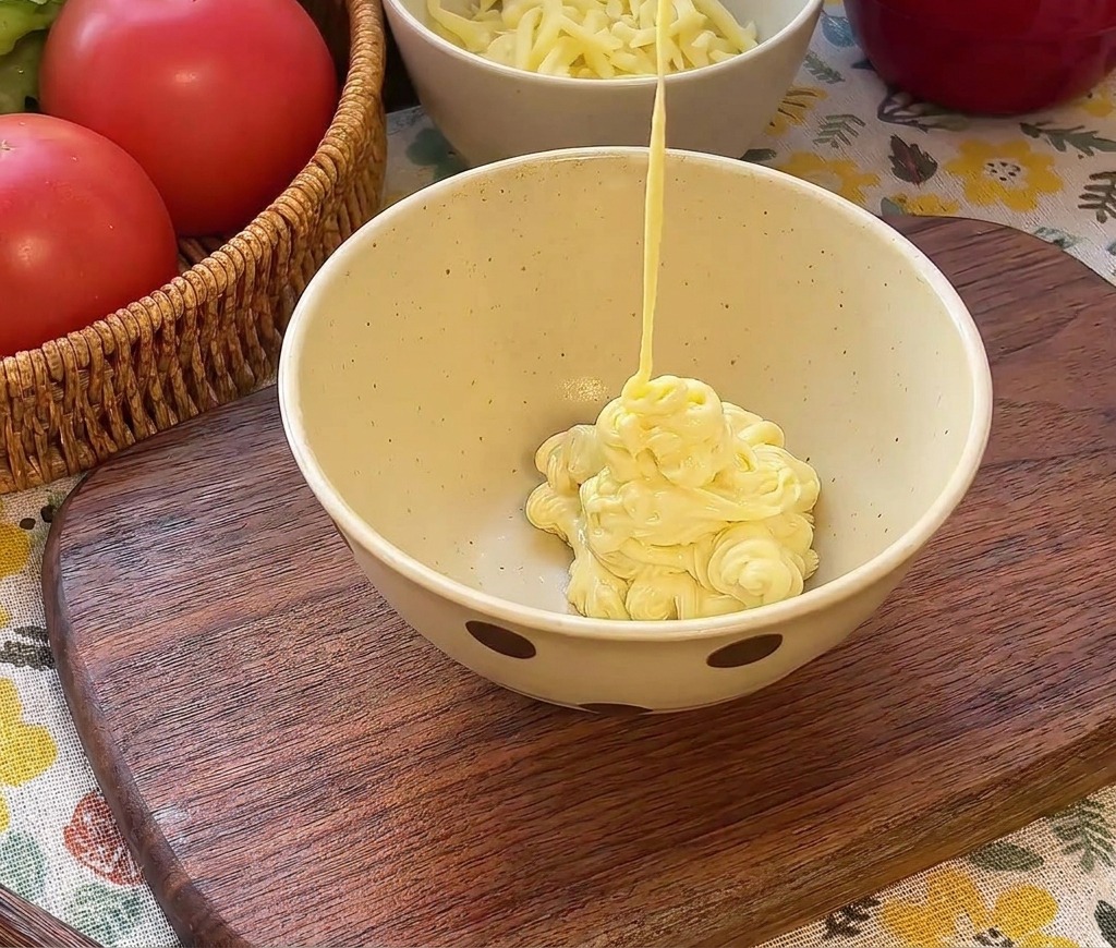 Creamy mayonnaise being poured into a bowl over red ketchup and green chopped pickles.