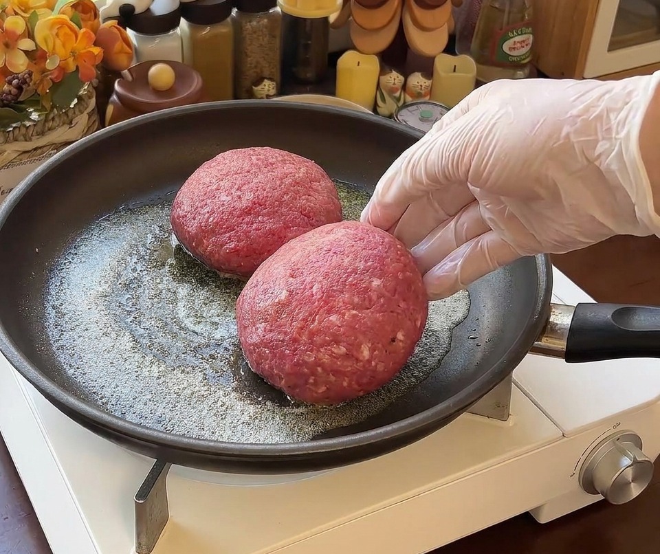 Two thick, round raw beef patties being placed into a frying pan with melted butter, with a gloved hand visible on the right.