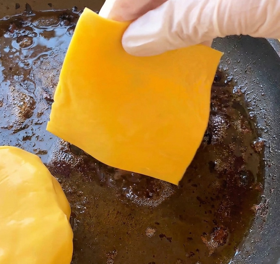 A gloved hand placing a bright yellow cheddar cheese slice onto a cooked burger patty sizzling in a pan.