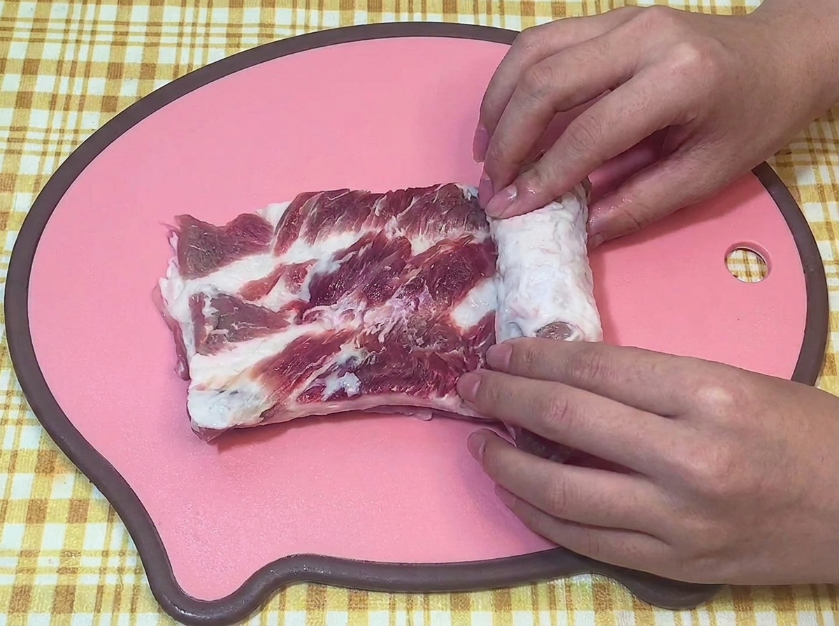 Hands tightly rolling a raw piece of pork belly into a uniform cylinder on a pink cutting board.