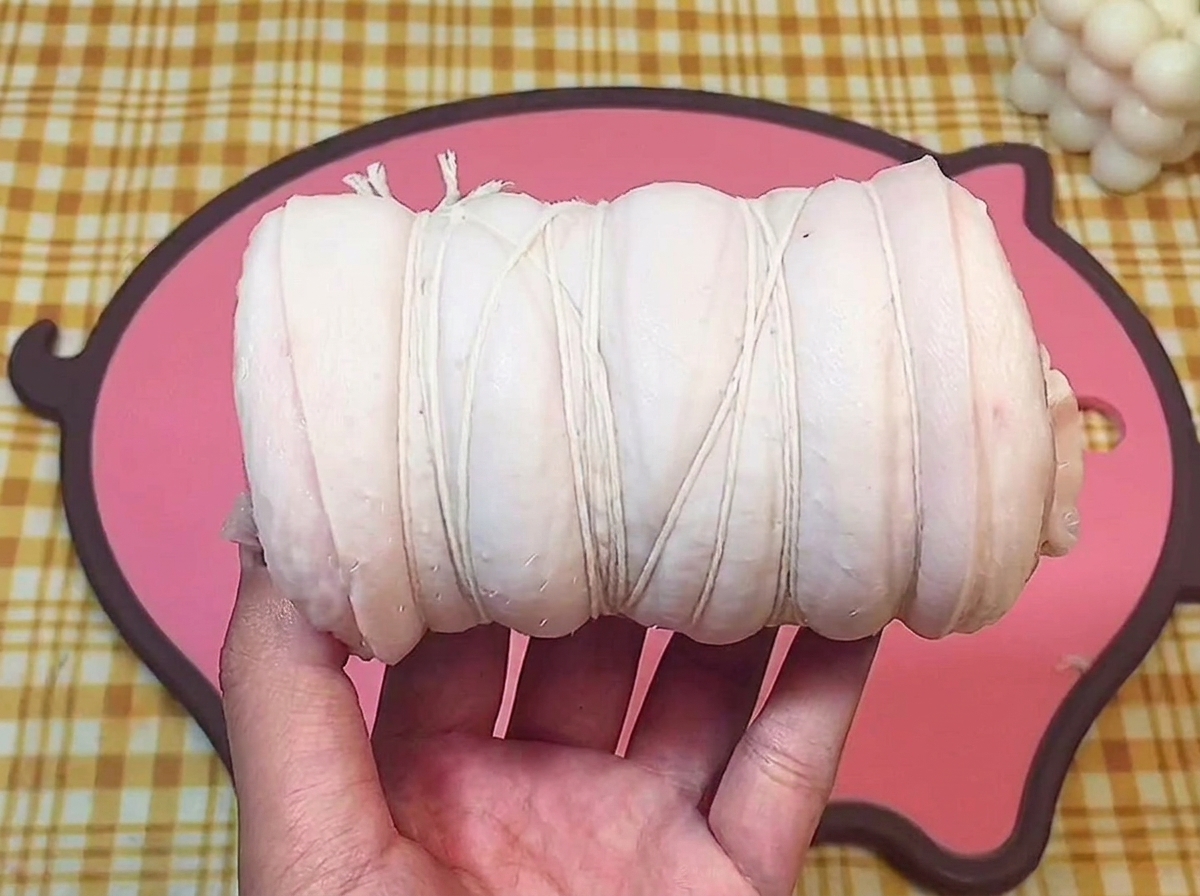 A hand holding a piece of raw pork belly tightly rolled and secured with white cooking twine over a pink cutting board.