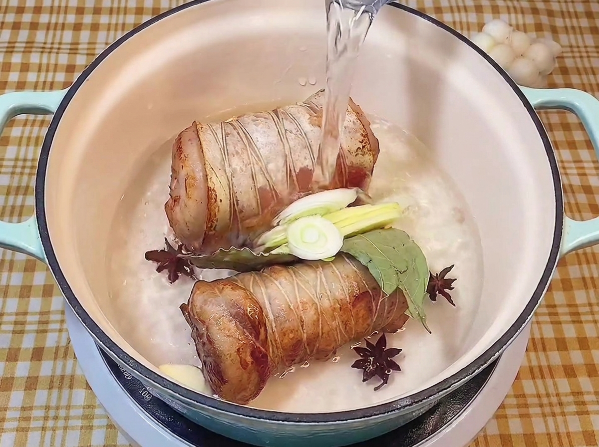 Pouring clear water into a cooking pot containing pan-fried pork belly rolls, aromatics, and spices.