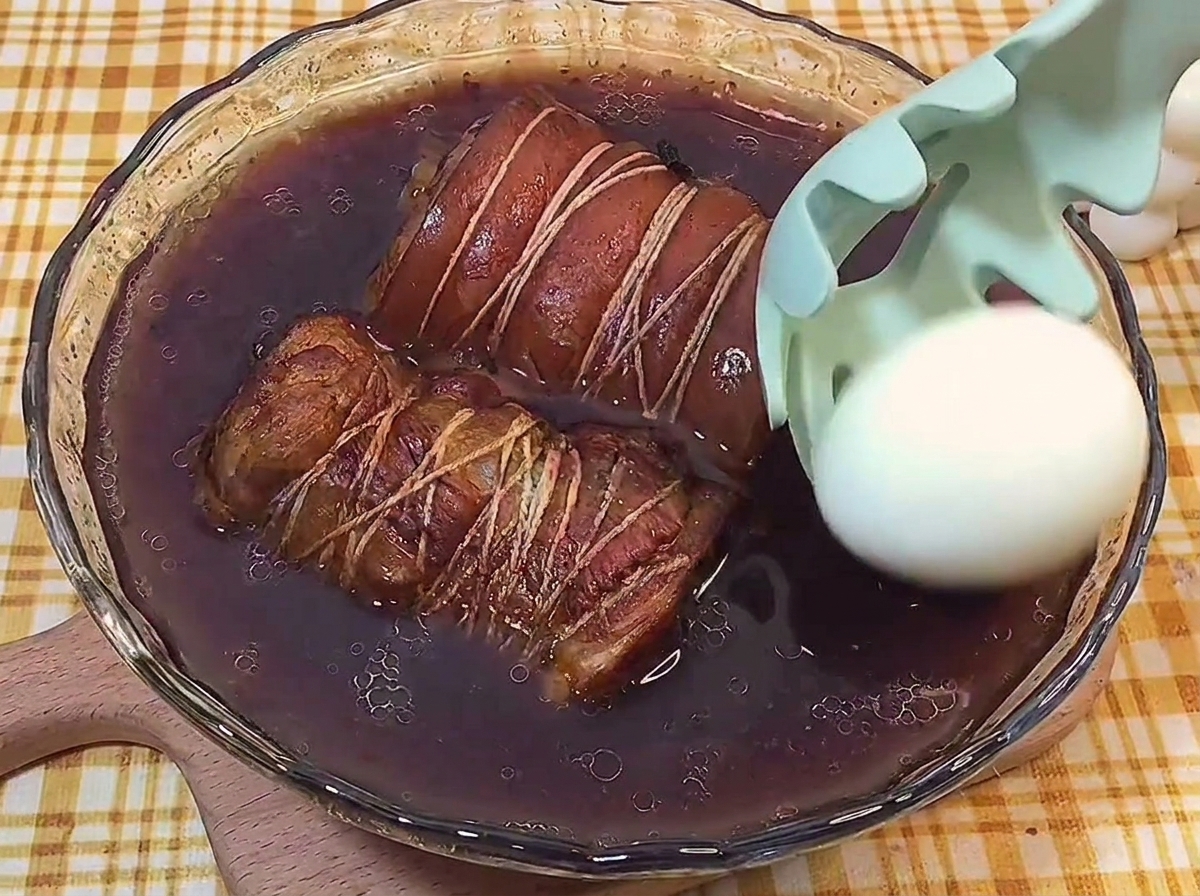 A peeled hard-boiled egg being placed with a slotted spoon into a glass dish filled with dark braising liquid and two tied rolls of pork belly.