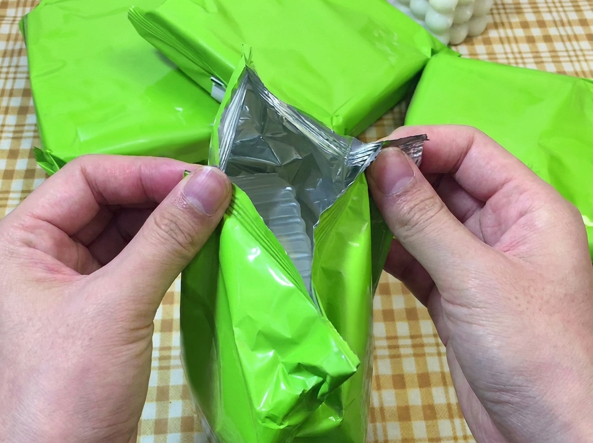 Two hands tearing open a bright green packet of instant ramen, exposing the plastic tray inside.