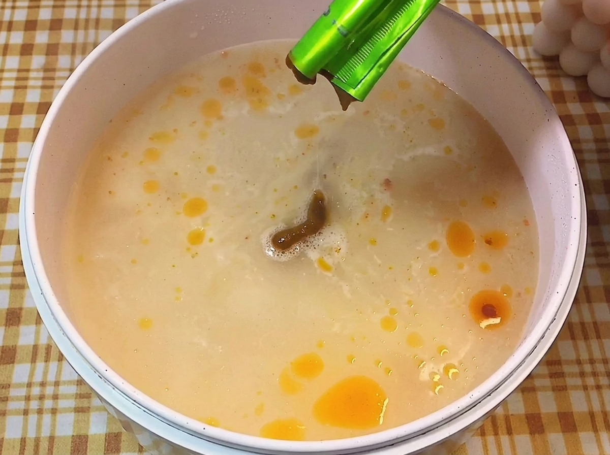Brown seasoning paste being squeezed from a green packet into a pot of simmering, milky broth dotted with oil.