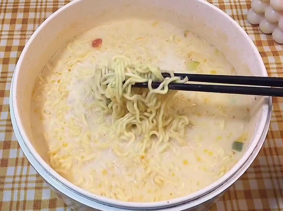 Black chopsticks pulling a portion of cooked wavy ramen noodles out of a creamy milky broth.