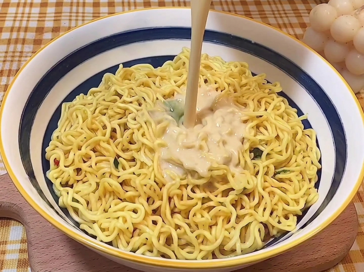 A stream of creamy, rich tonkotsu broth being poured over a bed of cooked ramen noodles in a serving bowl.