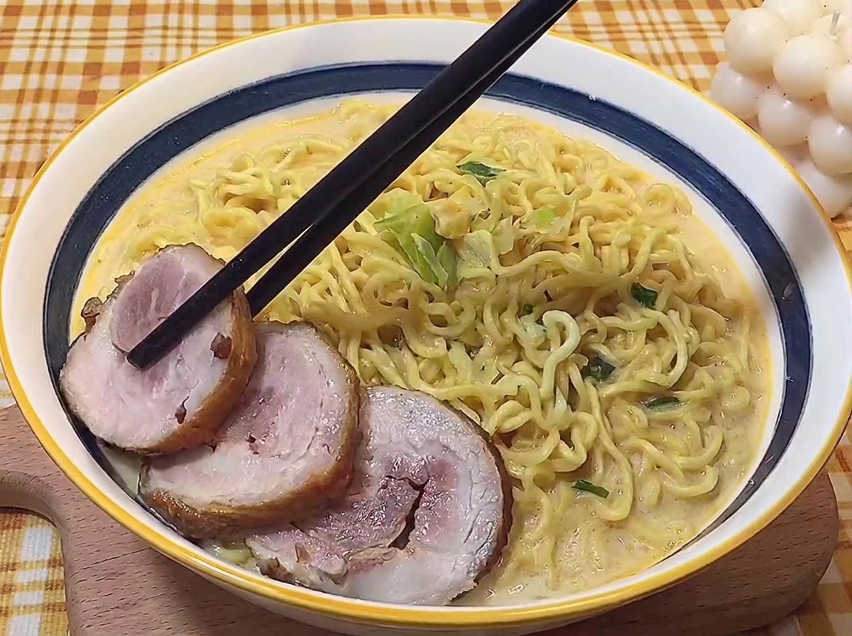 Chopsticks carefully placing slices of braised rolled pork belly into a bowl of hot tonkotsu ramen.
