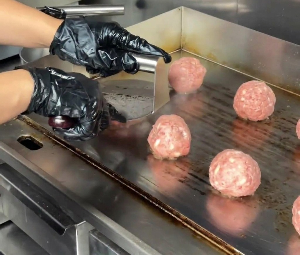A close up of a stainless steel burger press flattening a beef ball into a thick patty on a commercial flat top griddle.