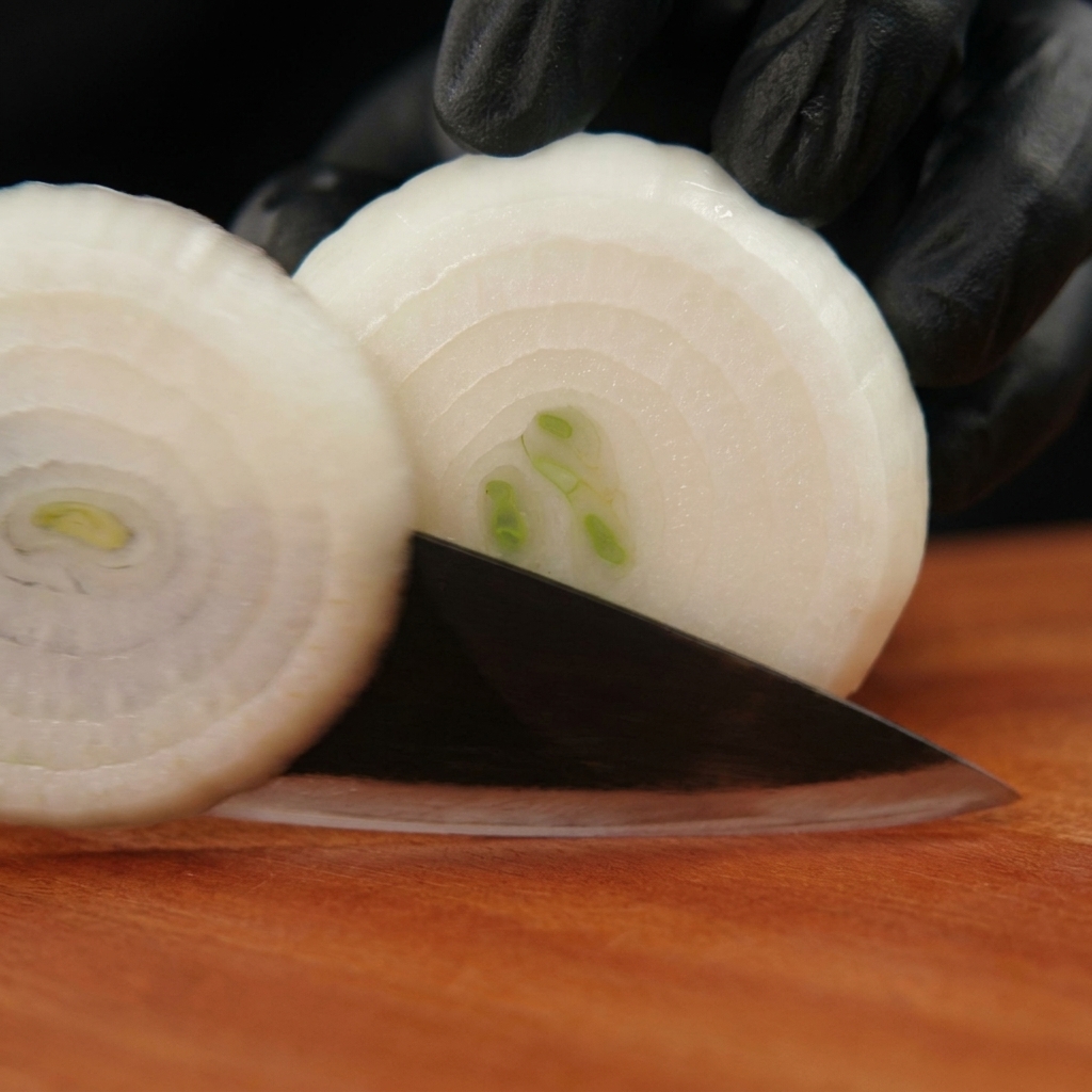 Black-gloved hands using a knife to cut a peeled white onion into rings on a wooden board.