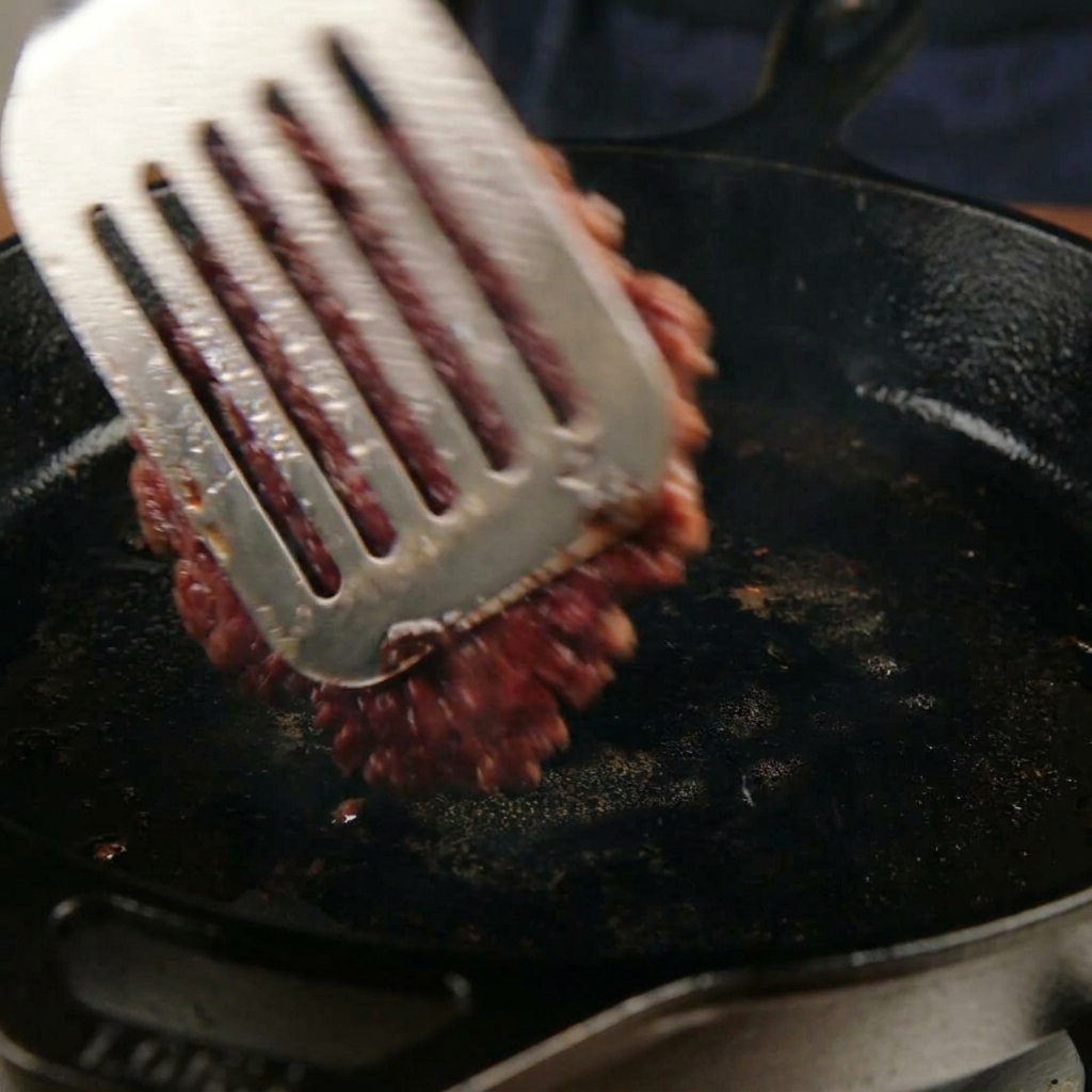 Flipping a caramelized, smashed beef patty in a skillet with a metal spatula.