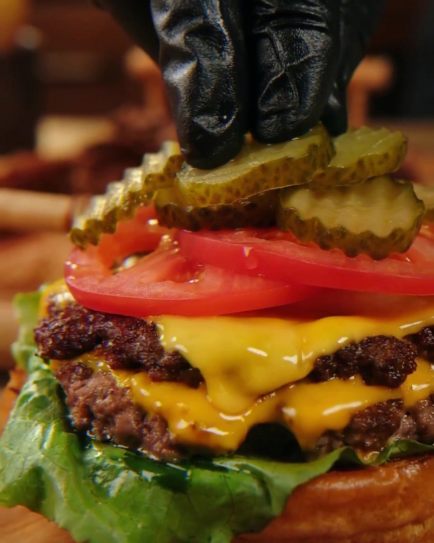 Black-gloved fingers carefully placing crinkle-cut pickle slices onto bright red tomato slices atop a stacked cheeseburger.