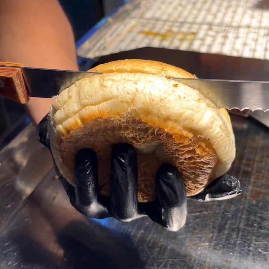 A person using a serrated knife to cut a large, soft burger bun in half horizontally on a stainless steel surface.