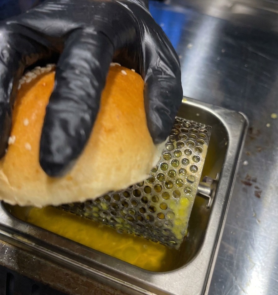 A gloved hand rolling the internal side of a burger bun over a butter roller in a metal tray.