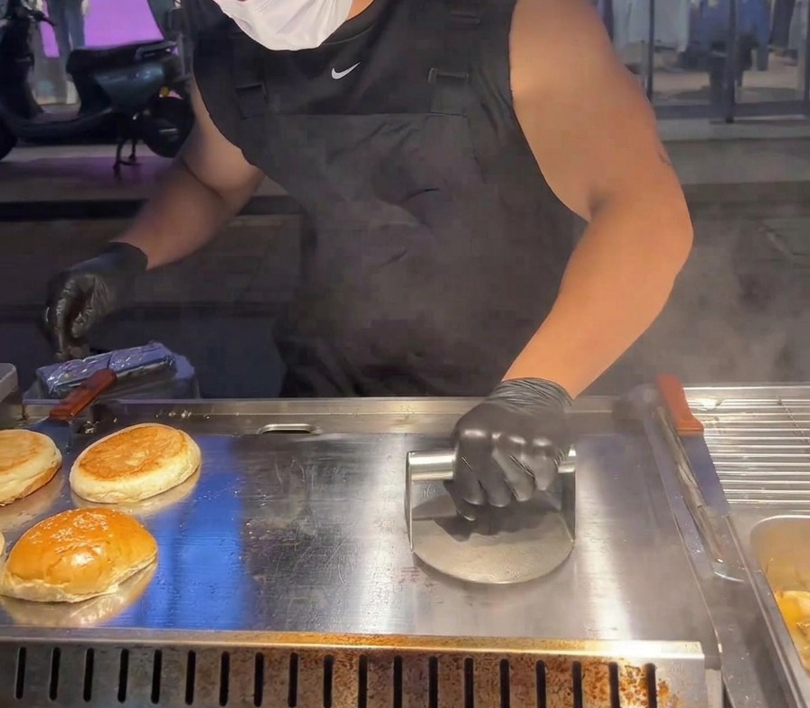 A circular heavy-duty metal press being used to flatten a beef patty against a searing hot griddle.