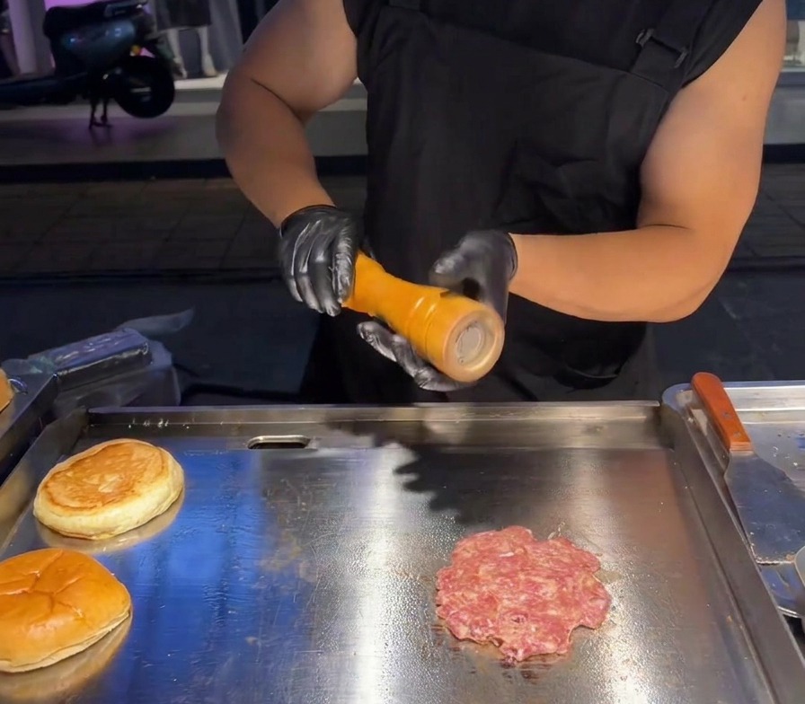 Fresh sea salt being ground from a mill onto the top of a flattened beef patty on a professional griddle.