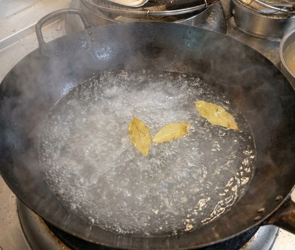 A large black wok filled with boiling water, with three bay leaves floating on the bubbling surface.