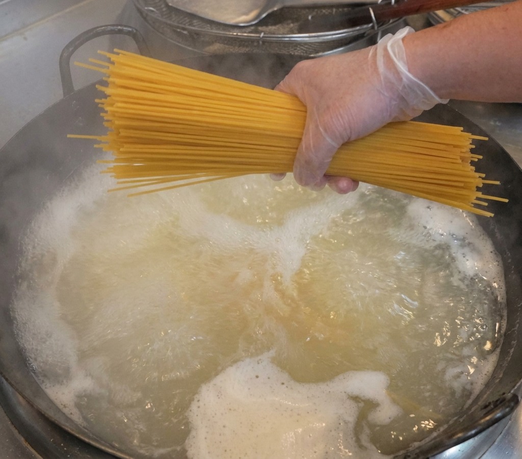 A hand wearing a clear glove holding a thick bundle of dry spaghetti over a pot of steaming, boiling water.