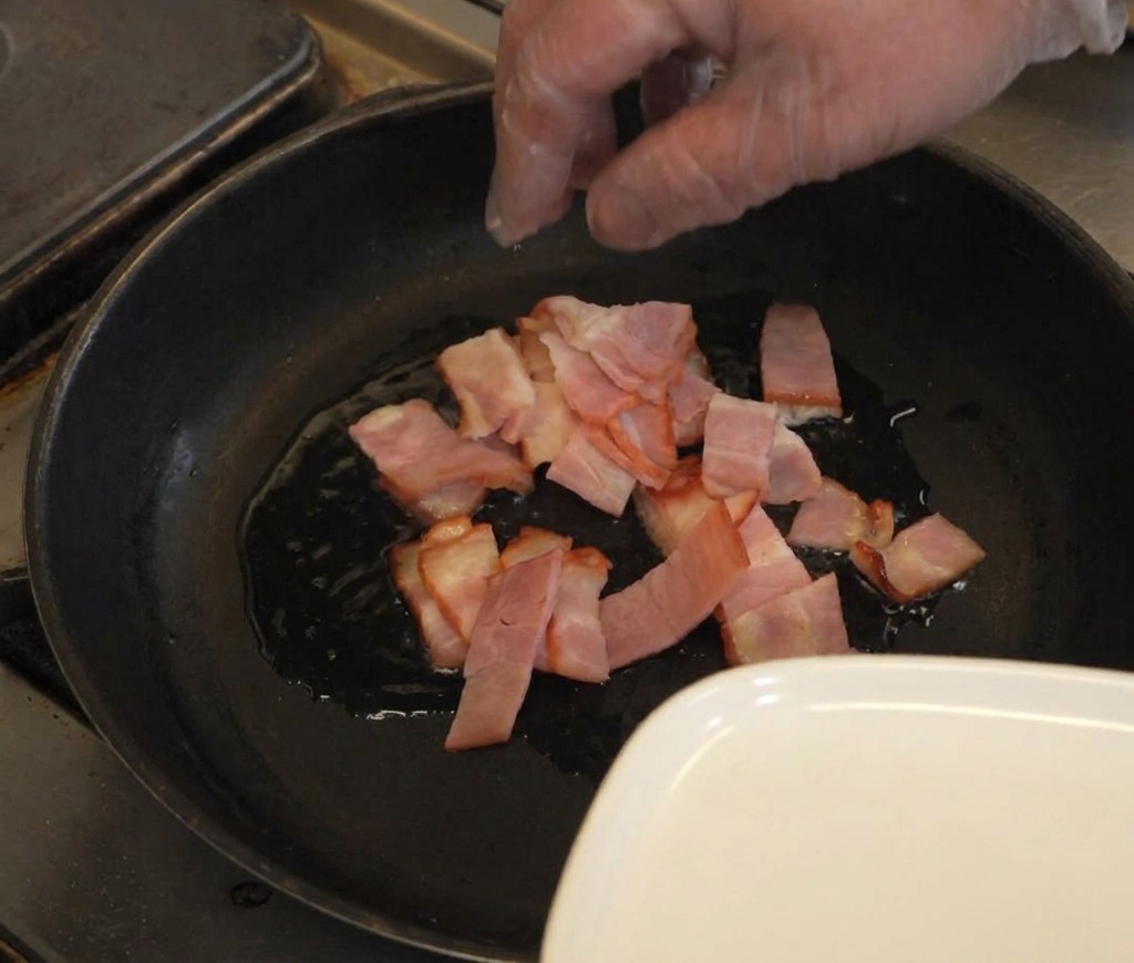 A hand placing sliced pieces of raw bacon into a dark frying pan with a thin layer of shimmering oil.