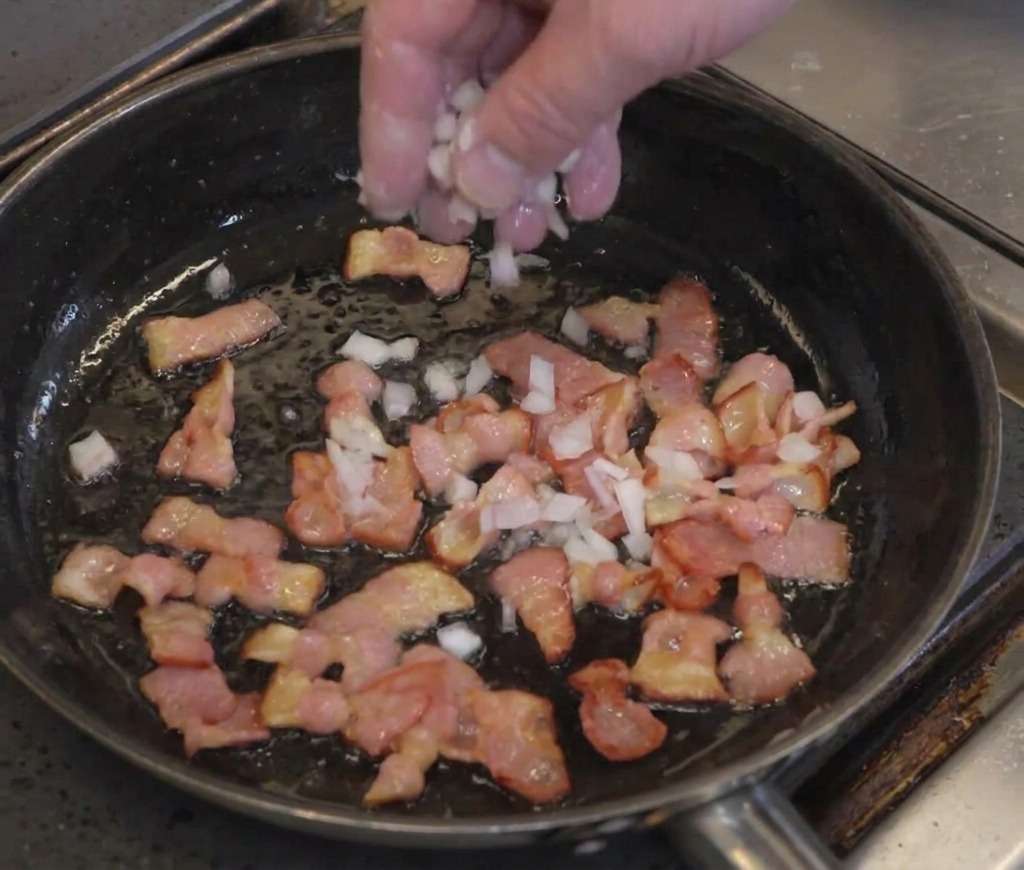 A hand sprinkling minced white onion over partially cooked bacon strips in a frying pan.