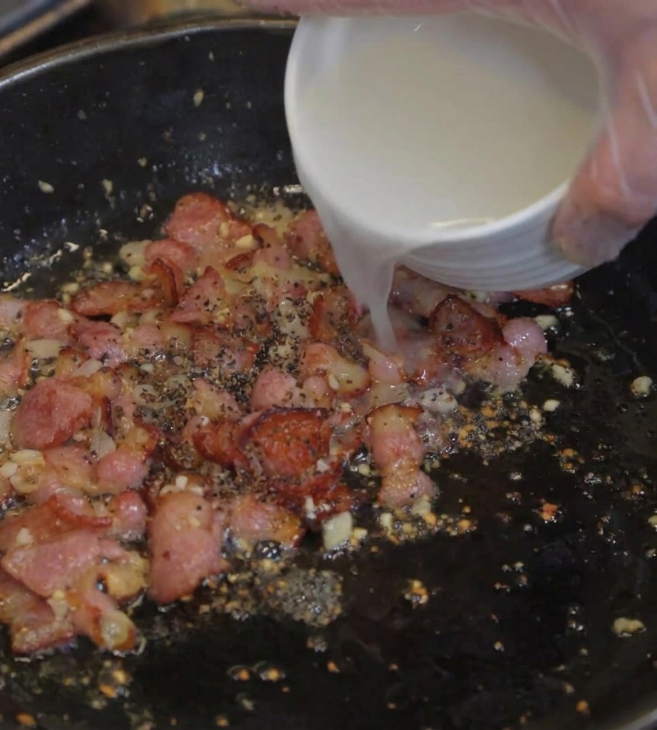 Cloudy pasta water being poured from a white bowl into a skillet containing crispy bacon and sautéed aromatics.