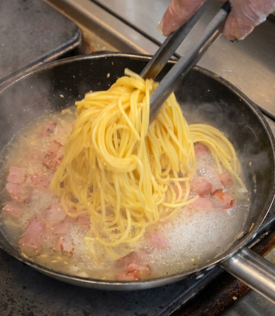A bundle of yellow spaghetti being lifted by tongs and placed into a pan with bacon bits and simmering liquid.