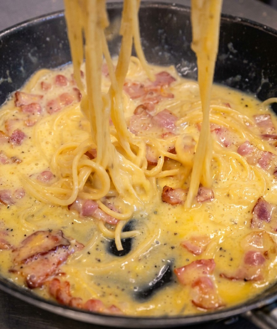 Spaghetti being tossed in a skillet, showing the sauce transforming into a thick, creamy, pale-yellow coating over the bacon and pasta.