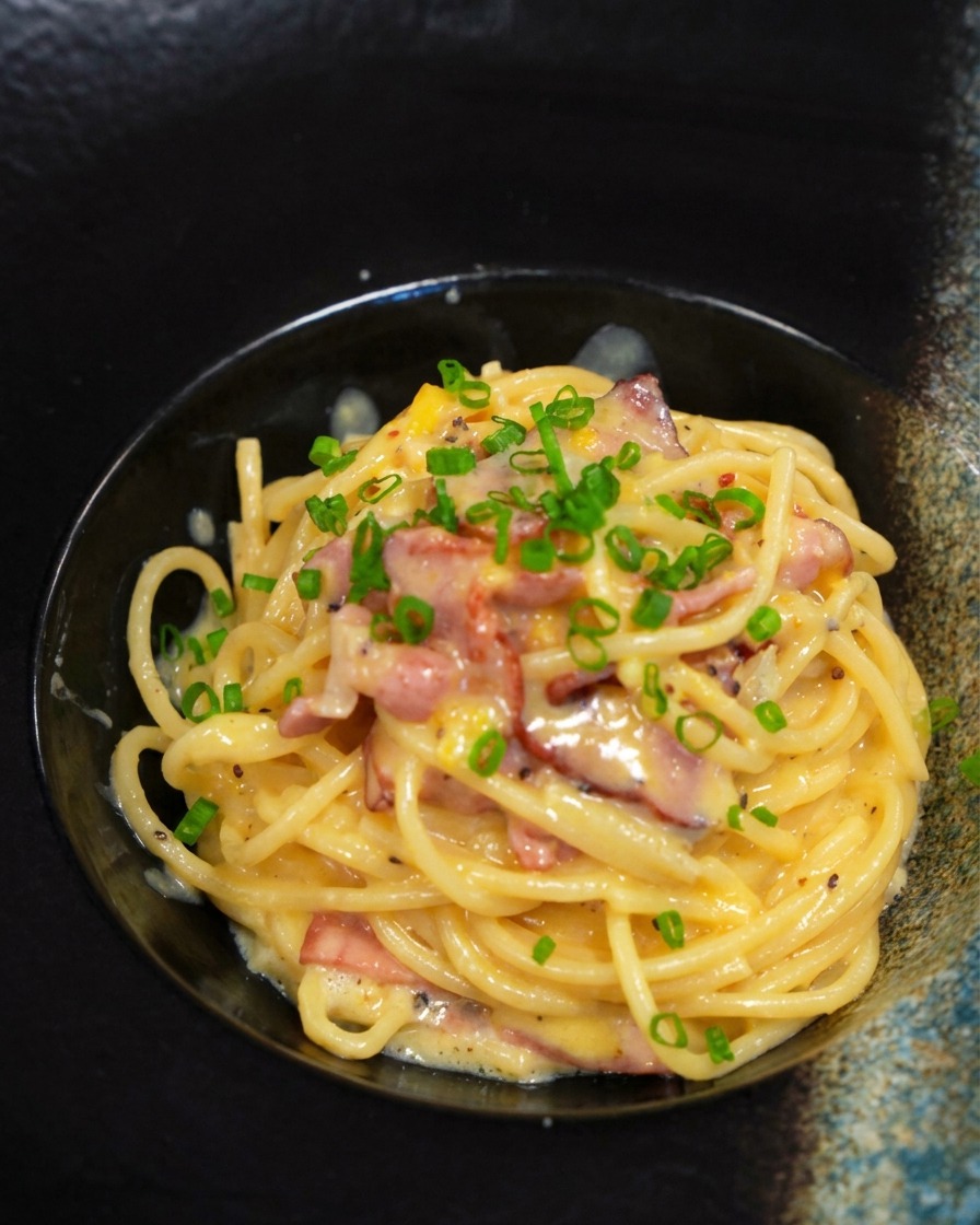 A finished serving of bacon carbonara in a black bowl being topped with a sprinkle of bright green chopped chives.