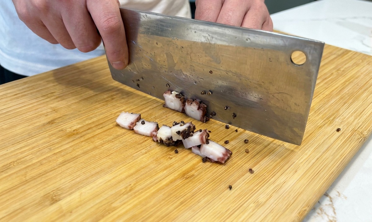 Slicing cured pork jowl into small cubes with a cleaver on a wooden board.