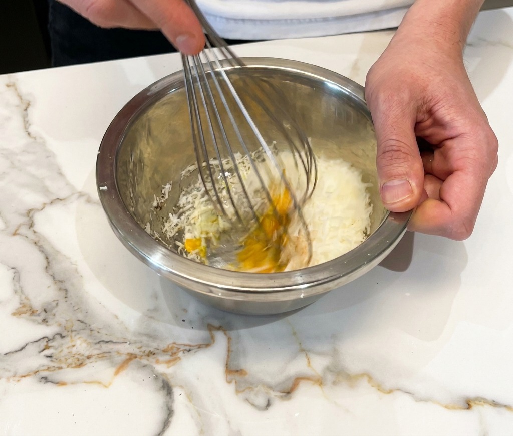 Whisking an egg, grated cheese, and black pepper together into a thick paste in a metal bowl.